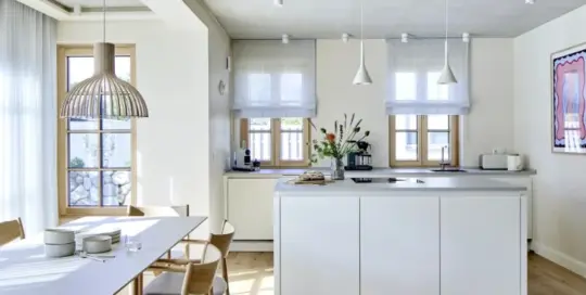 Küchenstudio Moderne Küche mit zentraler Insel, hellem Holzfußboden, Esstisch mit Stühlen, großen Fenstern und minimalistischem weißen Dekor.
