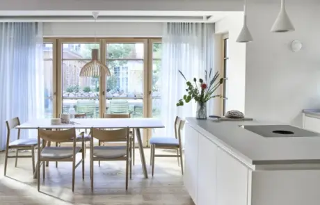 Küchenstudio Eine helle, moderne Küche und ein Essbereich mit hellen Holzmöbeln, weißen Schränken, Pendelleuchten und großen Fenstern mit transparenten Vorhängen mit Blick auf einen Garten.