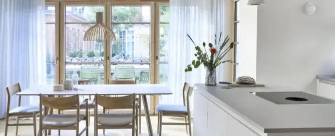 Küchenstudio Eine helle, moderne Küche und ein Essbereich mit hellen Holzmöbeln, weißen Schränken, Pendelleuchten und großen Fenstern mit transparenten Vorhängen mit Blick auf einen Garten.