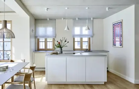 Küchenstudio Moderne Küche mit weißen Schränken, Insel und Pendelleuchten. Transparente Vorhänge verdecken die Fenster, und links ist ein Essbereich mit Holzstühlen zu sehen. An der rechten Wand hängt ein Kunstwerk.