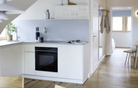 Küchenstudio Moderne weiße Küche mit minimalem Dekor, eingebautem Ofen, Geschirr und offenen Regalen; der angrenzende Flur führt zu einem hellen Zimmer mit Holzboden und einfacher Einrichtung.