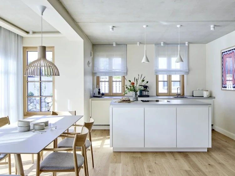 Küchenstudio Moderne Küche mit zentraler Insel, hellem Holzfußboden, Esstisch mit Stühlen, großen Fenstern und minimalistischem weißen Dekor.