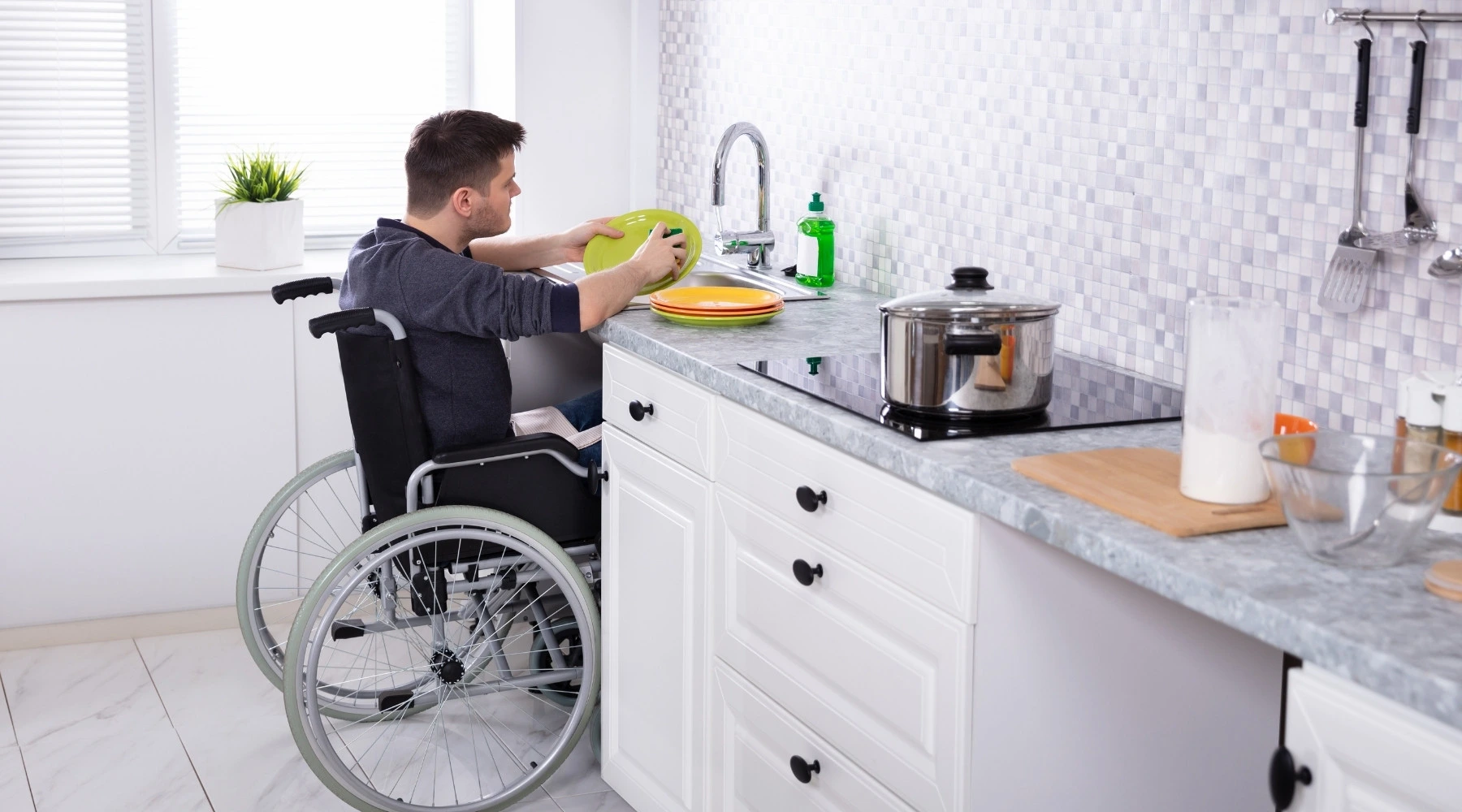 Küchenstudio Ein Mann im Rollstuhl wäscht sein Geschirr an einer Spüle ab, auf der saubere Teller, ein Topf auf dem Herd und ein Schneidebrett auf der Theke stehen.