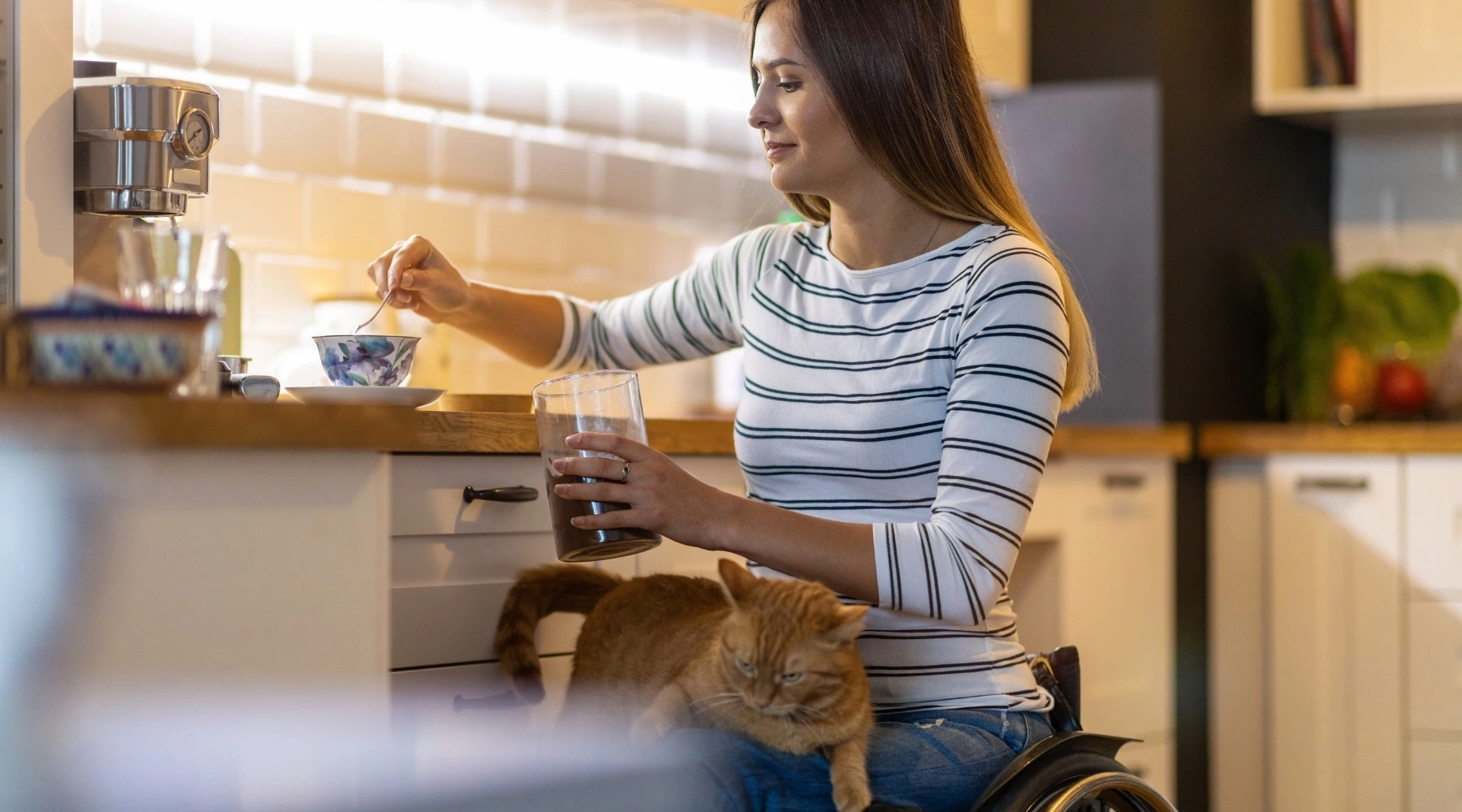 Küchenstudio Eine Frau im Rollstuhl sitzt in einer Küche, rührt mit der einen Hand in einer Schüssel und hält in der anderen ein Glas, während eine rothaarige Katze auf ihrem Schoß sitzt.