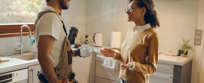 Küchenstudio Ein Handwerker in Arbeitskleidung unterhält sich mit einer Frau in einem legeren Pullover und einer Brille in einer modernen Küche.