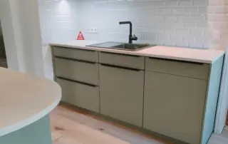 Küchenstudio Moderne Küche mit hellgrünen Schränken, schwarzer Spüle und Wasserhahn, weißer Arbeitsplatte und weißem Subway-Fliesenaufsatz; Holzboden und minimalistisches Design. Küchenstudio Moderne Küche mit hellgrünen Schränken, schwarzer Spüle und Wasserhahn, weißer Arbeitsplatte und weißem Subway-Fliesenaufsatz; Holzboden und minimalistisches Design.