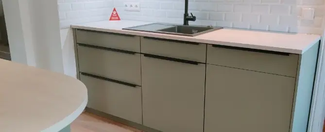 Küchenstudio Moderne Küche mit hellgrünen Schränken, schwarzer Spüle und Wasserhahn, weißer Arbeitsplatte und weißem Subway-Fliesenaufsatz; Holzboden und minimalistisches Design.