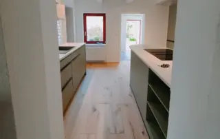 Küchenstudio Moderne Küche mit hellem Holzfußboden, zwei parallelen Arbeitsflächen, Einbaugeräten, offenen Regalen und einem rot gerahmten Fenster am Ende des Raumes. Küchenstudio Moderne Küche mit hellem Holzfußboden, zwei parallelen Arbeitsflächen, Einbaugeräten, offenen Regalen und einem rot gerahmten Fenster am Ende des Raumes.