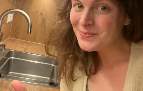 Küchenstudio Eine Frau mit langen braunen Haaren lächelt und zeigt den Daumen nach oben vor einer sauberen, leeren Küchenspüle in hellem Licht.