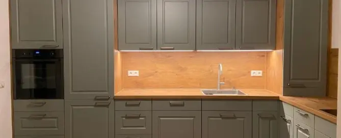 Küchenstudio Moderne Küche mit grauen Schränken, Arbeitsflächen und Aufkantung aus Holz, eingebautem Backofen, Spüle, Unterschrankbeleuchtung und einer Katze, die in der unteren linken Ecke auf dem Boden sitzt.