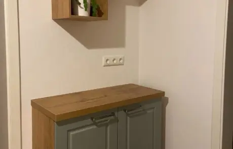 Küchenstudio Holzschrank mit grünen Türen, darunter ein passendes Wandregal mit einer Topfpflanze, vor einer schlichten hellen Wand mit drei Steckdosen.