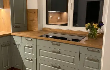 Küchenstudio Moderne Küche mit hellgrünen Schränken, Arbeitsplatten aus Holz, einem Elektrokochfeld unter einem Fenster und einer Blumenvase auf dem Tresen.