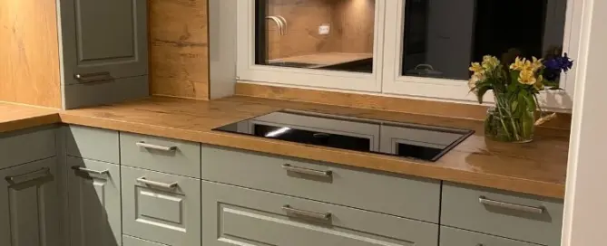 Küchenstudio Moderne Küche mit hellgrünen Schränken, Arbeitsplatten aus Holz, einem Elektrokochfeld unter einem Fenster und einer Blumenvase auf dem Tresen.