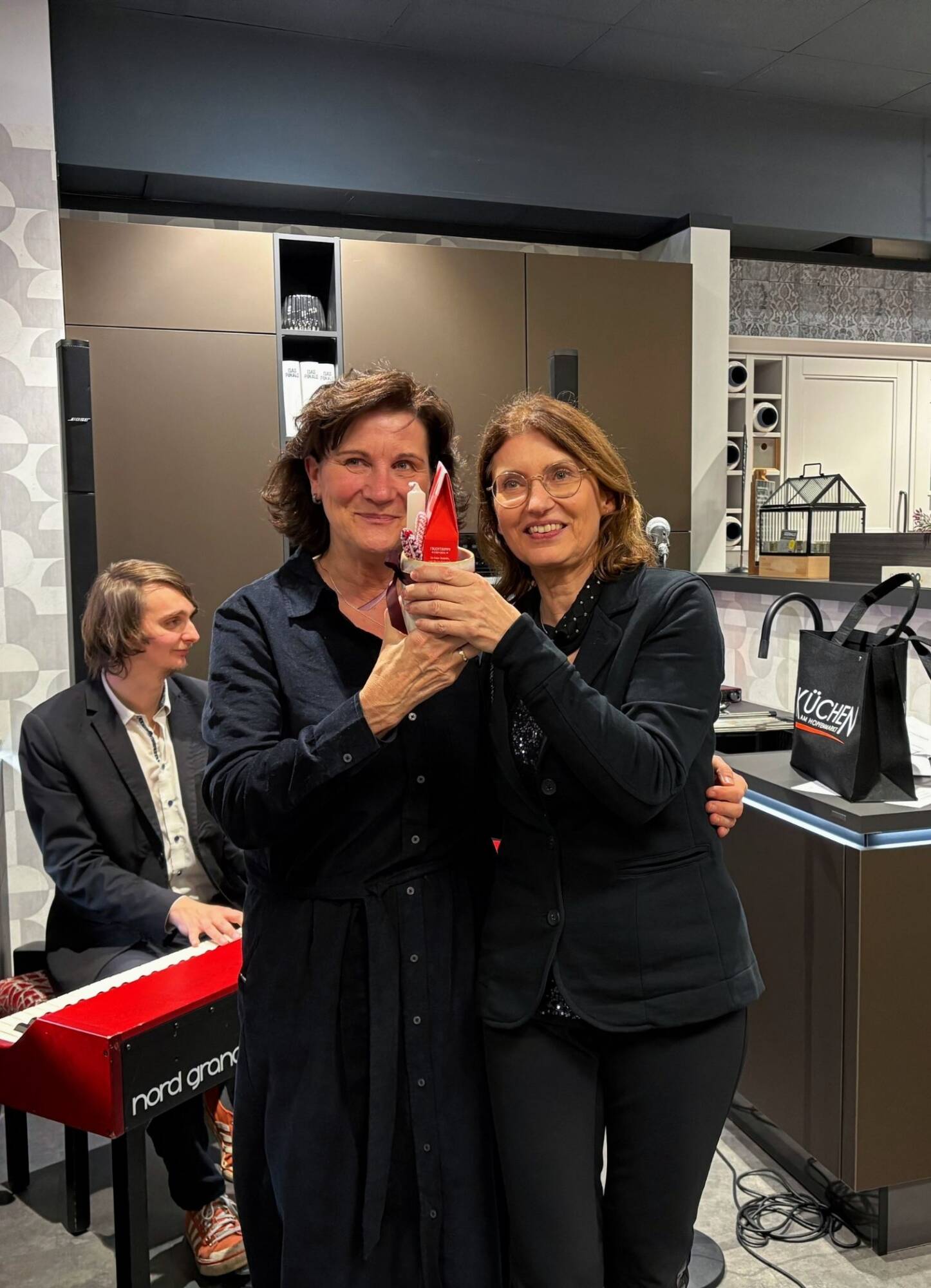 Küchenstudio Zwei Frauen stehen zusammen und halten einen roten Preis in einer modernen Küche, während ein Mann im Hintergrund auf einem Keyboard spielt.