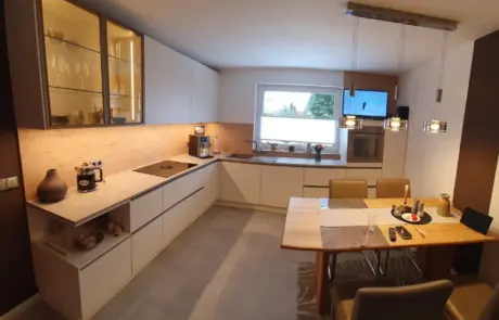 Küchenstudio Moderne Küche mit weißen Schränken, Einbaugeräten, einer Kaffeemaschine und einem Esstisch für zwei Personen unter Hängelampen. Ein Fenster sorgt für natürliches Licht über dem Spülbereich.