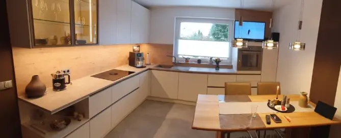Küchenstudio Moderne Küche mit weißen Schränken, Einbaugeräten, einer Kaffeemaschine und einem Esstisch für zwei Personen unter Hängelampen. Ein Fenster sorgt für natürliches Licht über dem Spülbereich.