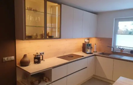 Küchenstudio Moderne Küche mit weißen Schränken, oberen Glasfächern, eingebautem Kochfeld, Kaffeemaschine und einem Fenster über der Spüle; neutrale Farbtöne und Unterschrankbeleuchtung.