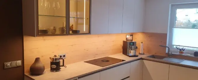Küchenstudio Moderne Küche mit weißen Schränken, oberen Glasfächern, eingebautem Kochfeld, Kaffeemaschine und einem Fenster über der Spüle; neutrale Farbtöne und Unterschrankbeleuchtung.