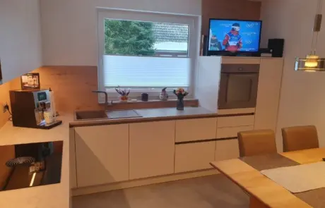 Küchenstudio Moderne Küche mit weißen Schränken, Einbaugeräten, einer Kaffeemaschine und einem Esstisch; ein Fernseher auf dem Tresen zeigt eine Wintersportübertragung.