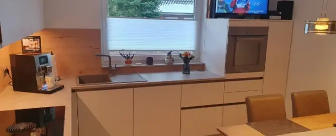 Küchenstudio Moderne Küche mit weißen Schränken, Einbaugeräten, einer Kaffeemaschine und einem Esstisch; ein Fernseher auf dem Tresen zeigt eine Wintersportübertragung.