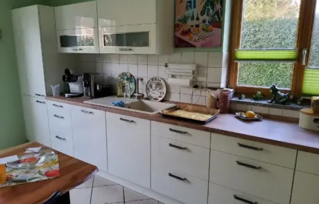 Küchenstudio Moderne Küche mit weißen Schränken, einer Arbeitsplatte aus Holz, Kaffeemaschinen, Geschirr und einem Tablett mit Teig am Spülbecken. Ein Fenster mit grünen Jalousien lässt natürliches Licht herein.