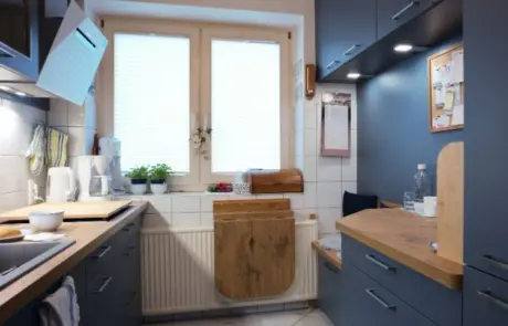 Küchenstudio Eine kleine Küche mit blauen Schränken, Arbeitsflächen aus Holz, einem Fenster mit Jalousien, Topfpflanzen und einem klappbaren Holztisch, der unter dem Fenster angebracht ist.