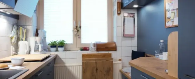 Küchenstudio Eine kleine Küche mit blauen Schränken, Arbeitsflächen aus Holz, einem Fenster mit Jalousien, Topfpflanzen und einem klappbaren Holztisch, der unter dem Fenster angebracht ist.