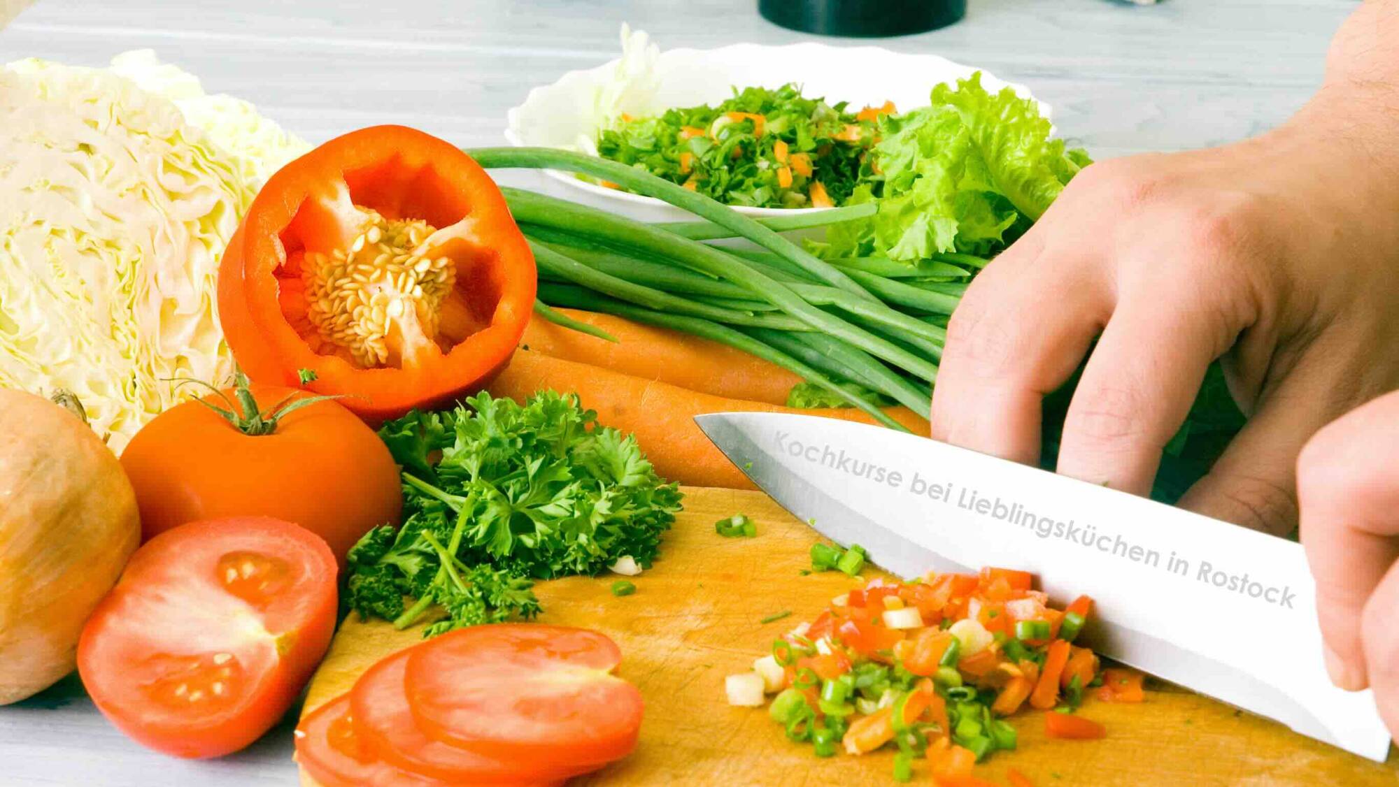 Küchenstudio Eine Person schneidet Gemüse auf einem Schneidebrett, umgeben von Tomaten, Paprika, Karotten, Kohl, Petersilie und grünen Zwiebeln, und bereitet gesunde Mahlzeiten für einen Kochabend für Frauen mit Krebs vor. Auf der Klinge des Messers steht ein deutscher Text.