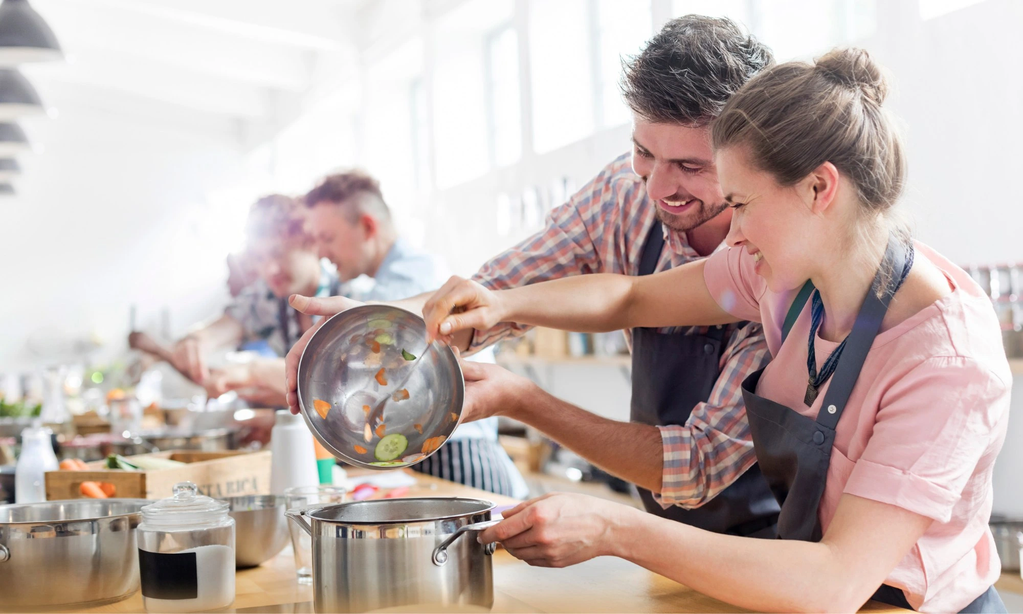 Küchenstudio Zwei Personen in Schürzen lächeln, während sie in einem hellen Küchenstudio gehacktes Gemüse aus einer Rührschüssel in einen Topf schütten, während andere im Hintergrund ihre Lieblingsküchen zubereiten. Küchenstudio Zwei Personen in Schürzen lächeln, während sie in einem hellen Küchenstudio gehacktes Gemüse aus einer Rührschüssel in einen Topf schütten, während andere im Hintergrund ihre Lieblingsküchen zubereiten.
