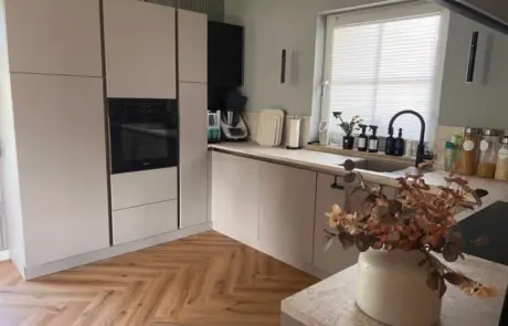 Küchenstudio Moderne Küche mit hellen Schränken, Einbaubackofen, Fischgrätenparkett, Küchengeräten auf der Arbeitsplatte, Topfpflanzen und Vorratsgläsern neben einem Fenster mit Jalousie. Küchenstudio Moderne Küche mit hellen Schränken, Einbaubackofen, Fischgrätenparkett, Küchengeräten auf der Arbeitsplatte, Topfpflanzen und Vorratsgläsern neben einem Fenster mit Jalousie.
