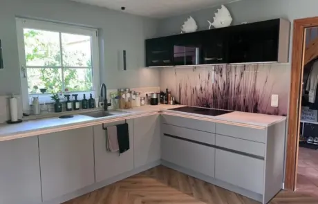 Küchenstudio Moderne Küche mit hellgrauen Schränken, einem Boden in Holzoptik, schwarzen Oberschränken und einer Küchenrückwand mit einem Motiv aus hohem Gras. Durch ein Fenster über der Spüle strömt Sonnenlicht herein. Küchenstudio Moderne Küche mit hellgrauen Schränken, einem Boden in Holzoptik, schwarzen Oberschränken und einer Küchenrückwand mit einem Motiv aus hohem Gras. Durch ein Fenster über der Spüle strömt Sonnenlicht herein.
