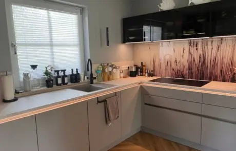 Küchenstudio Moderne Küche mit hellgrauen Schränken, schwarzen Arbeitsplattenelementen, einem Elektrokochfeld und einer dekorativen Fliesenwand mit Grasmotiv unter den schwarzen Hängeschränken. Durch ein Fenster fällt Tageslicht herein. Küchenstudio Moderne Küche mit hellgrauen Schränken, schwarzen Arbeitsplattenelementen, einem Elektrokochfeld und einer dekorativen Fliesenwand mit Grasmotiv unter den schwarzen Hängeschränken. Durch ein Fenster fällt Tageslicht herein.