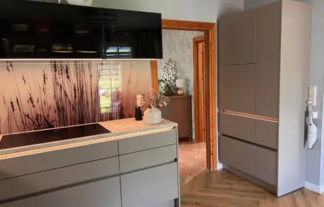 Küchenstudio Moderne Küche mit mattgrauen Schränken, schwarzen Glasoberschränken, einem Induktionskochfeld und einer Wandtapete mit hohem Grasmotiv; eine offene Tür führt in einen weiteren Raum mit ähnlicher Einrichtung. Küchenstudio Moderne Küche mit mattgrauen Schränken, schwarzen Glasoberschränken, einem Induktionskochfeld und einer Wandtapete mit hohem Grasmotiv; eine offene Tür führt in einen weiteren Raum mit ähnlicher Einrichtung.
