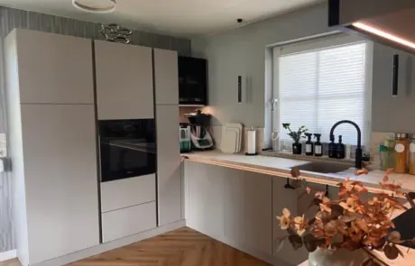 Küchenstudio Moderne Küche mit mattgrauen Schränken, Einbaubackofen, auf der Arbeitsplatte stehenden Küchengeräten, einem Fenster mit Jalousie, schwarzen Armaturen und einer Vase mit Trockenblumen im Vordergrund. Küchenstudio Moderne Küche mit mattgrauen Schränken, Einbaubackofen, auf der Arbeitsplatte stehenden Küchengeräten, einem Fenster mit Jalousie, schwarzen Armaturen und einer Vase mit Trockenblumen im Vordergrund.