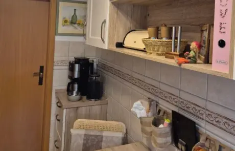 Küchenstudio Kleine Küchenecke mit einer Holztür, Küchengeräten auf einer Arbeitsplatte, Regalen mit Körben und Küchenutensilien, einem rosa Ordner sowie gefliesten Wänden mit einem Muster aus Beige- und Brauntönen.