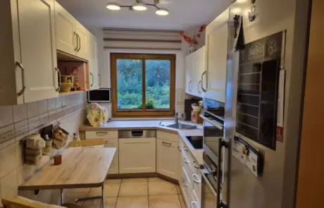 Küchenstudio Eine schmale Küche mit weißen Schränken, Edelstahlgeräten, einem Holztisch mit zwei Stühlen und einem Fenster mit Blick ins Grüne.