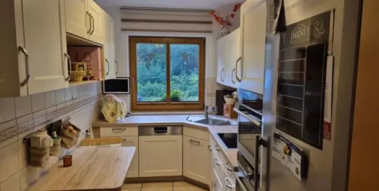 Küchenstudio Eine schmale Küche mit weißen Schränken, Edelstahlgeräten, einem Holztisch mit zwei Stühlen und einem Fenster mit Blick ins Grüne.